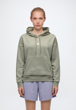 Un mannequin se tient de face, vêtu d’un sweat à capuche vert sauge et d’un short couleur lavande, sur un fond blanc uni.