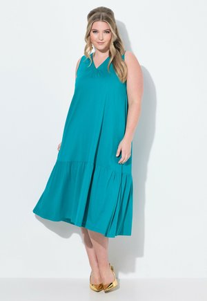 Tiefblaues, ärmelloses Kleid mit V-Ausschnitt und weit auslaufendem, gestuftem Rock. Aus weichem Stoff mit glatter Textur gefertigt. Kombiniert mit goldenen Absätzen.