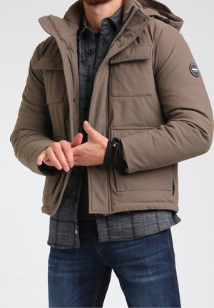 Bruine gewatteerde jas met een capuchon, voorzien van meerdere zakken aan de voorkant en een zwarte logopatch, gedragen over een donker geruit overhemd en donkere spijkerbroek.