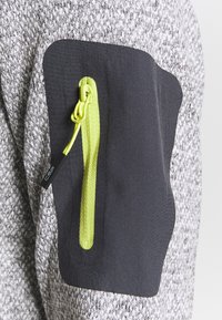 Grijs textiel met een zwarte patchzak, voorzien van een verticale neongele rits en trekband op een mouw van het kledingstuk.