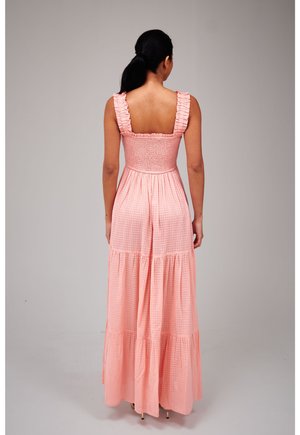 Kobieta o ciemnych włosach w różowej sukni maxi bez rękawów z falbankowymi ramiączkami i warstwową spódnicą, stojąca tyłem na jednolitym tle.