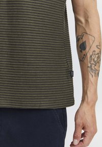 Camisa a rayas verde oliva con tejido texturizado, que presenta una etiqueta de logo en el lado; combinada con pantalones azul marino oscuros.