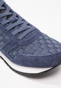 Gros plan d'une basket bleu marine avec tissu tissé, empiècements en daim, lacets assortis, semelle intermédiaire blanche et semelle extérieure en caoutchouc noir sur fond blanc.