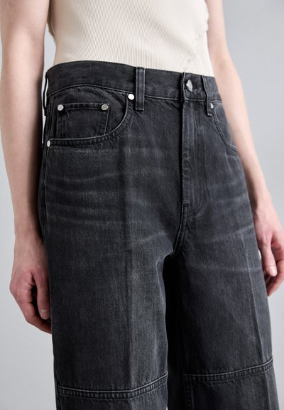 Jean en denim noir à taille haute, avec cinq poches et des éléments en métal argenté. Le tissu présente une texture délavée avec des coutures et des détails distincts.