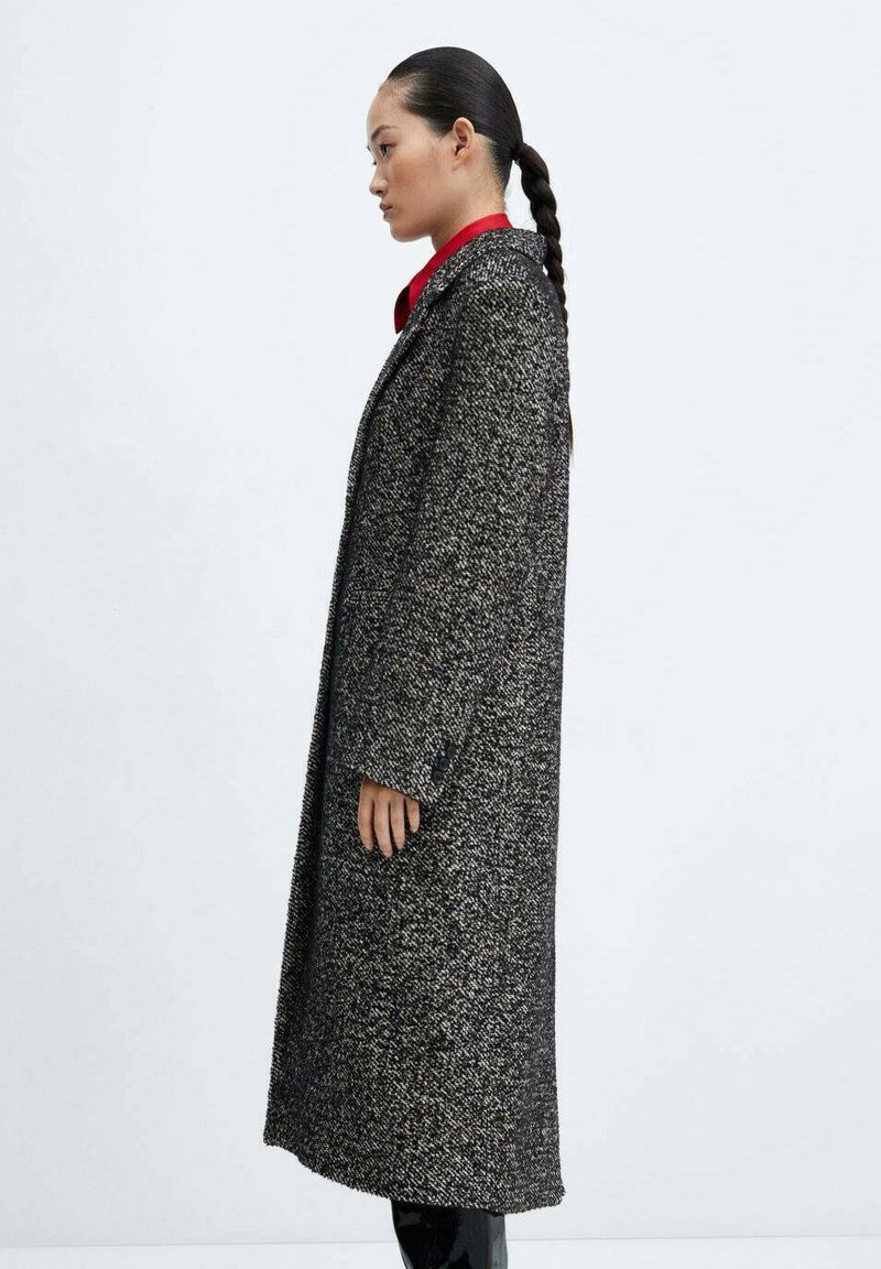 Lungo cappotto in tweed grigio e nero con colletto, tasche laterali, vestibilità dritta e tessuto strutturato, indossato sopra una camicia rossa.