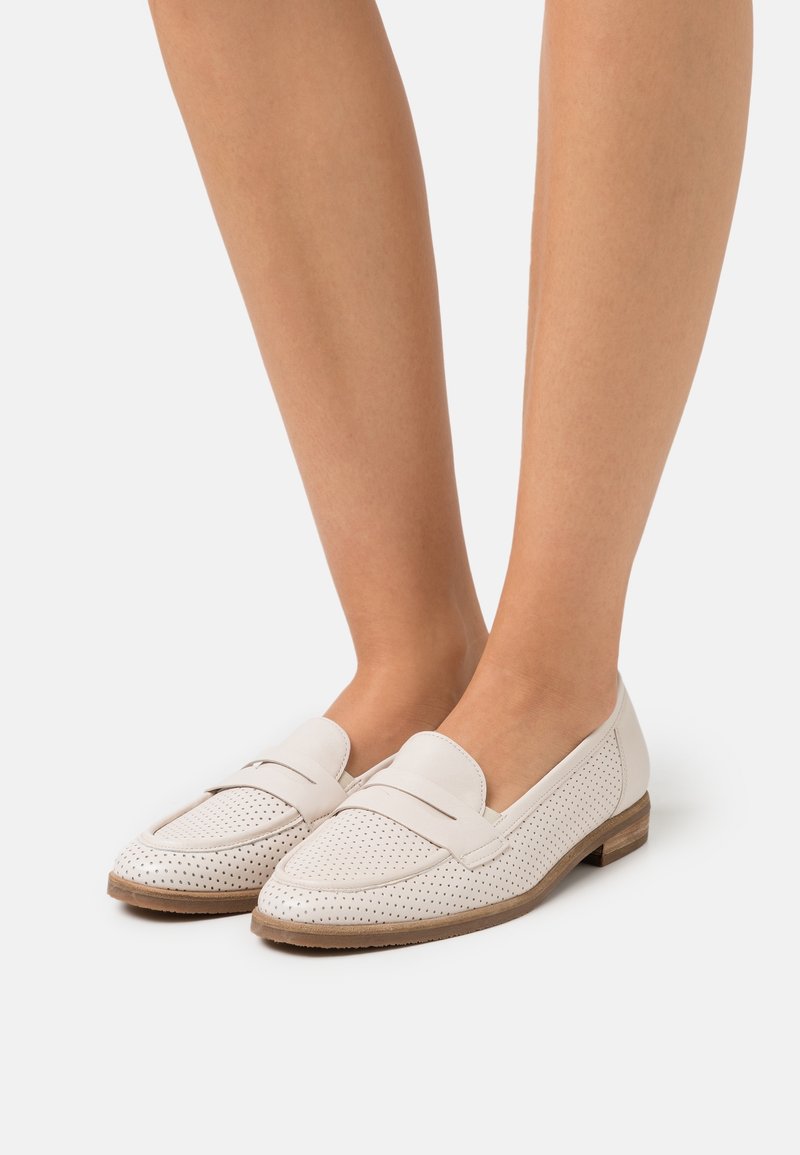 Pieds portant des mocassins en cuir blanc perforé avec un petit talon en bois, présentés sur un fond uni gris clair.