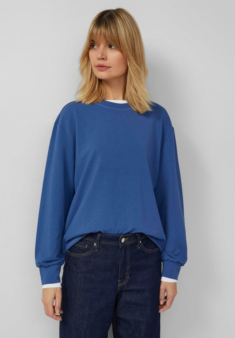 Donna che indossa una felpa blu a maniche lunghe sopra una camicia bianca e jeans blu scuro, in piedi contro uno sfondo grigio chiaro uniforme.