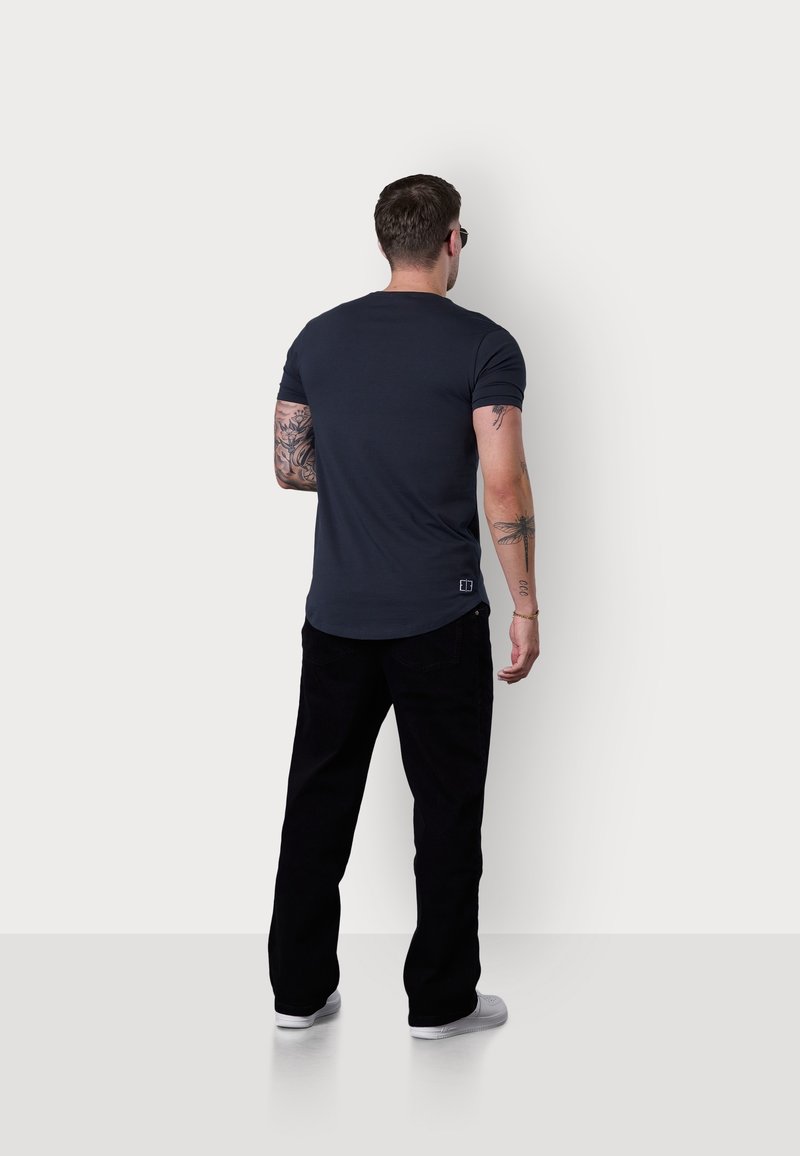 Homme avec des tatouages portant des lunettes de soleil noires, un t-shirt foncé, un pantalon noir et des chaussures blanches, debout face à un mur blanc uni.