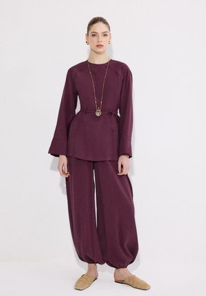 Γυναίκα με χαλαρή δαμασκηνή εμφάνιση με παντελόνι πλατύ και μπλούζα μακριών μανικιών, διακοσμημένη με μακρύ κολιέ με μενταγιόν και χρυσά σκουλαρίκια.