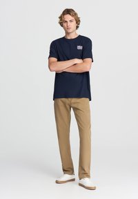T-shirt in cotone blu navy con maniche corte e grafica sul petto, abbinato a pantaloni marrone chiaro e sneaker bianche. Focus sul design casual.