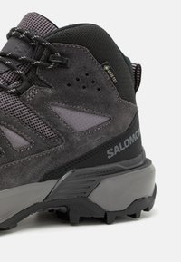 Grå tursko med semsket og mesh-materialer, med en robust såle, GORE-TEX-merke, avansert chassis og en distinkt teksturert design.