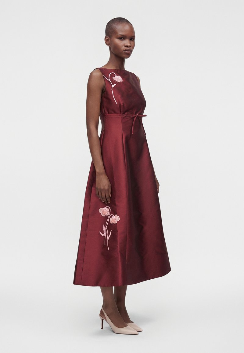 Femme debout dans une robe midi bordeaux sans manches avec broderie florale rose et talons beiges sur fond blanc uni.