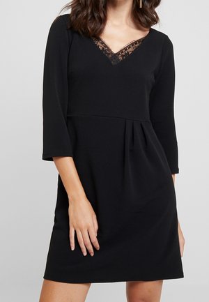 Mujer con vestido negro de manga tres cuartos y escote en V con encaje, sobre un fondo gris liso.