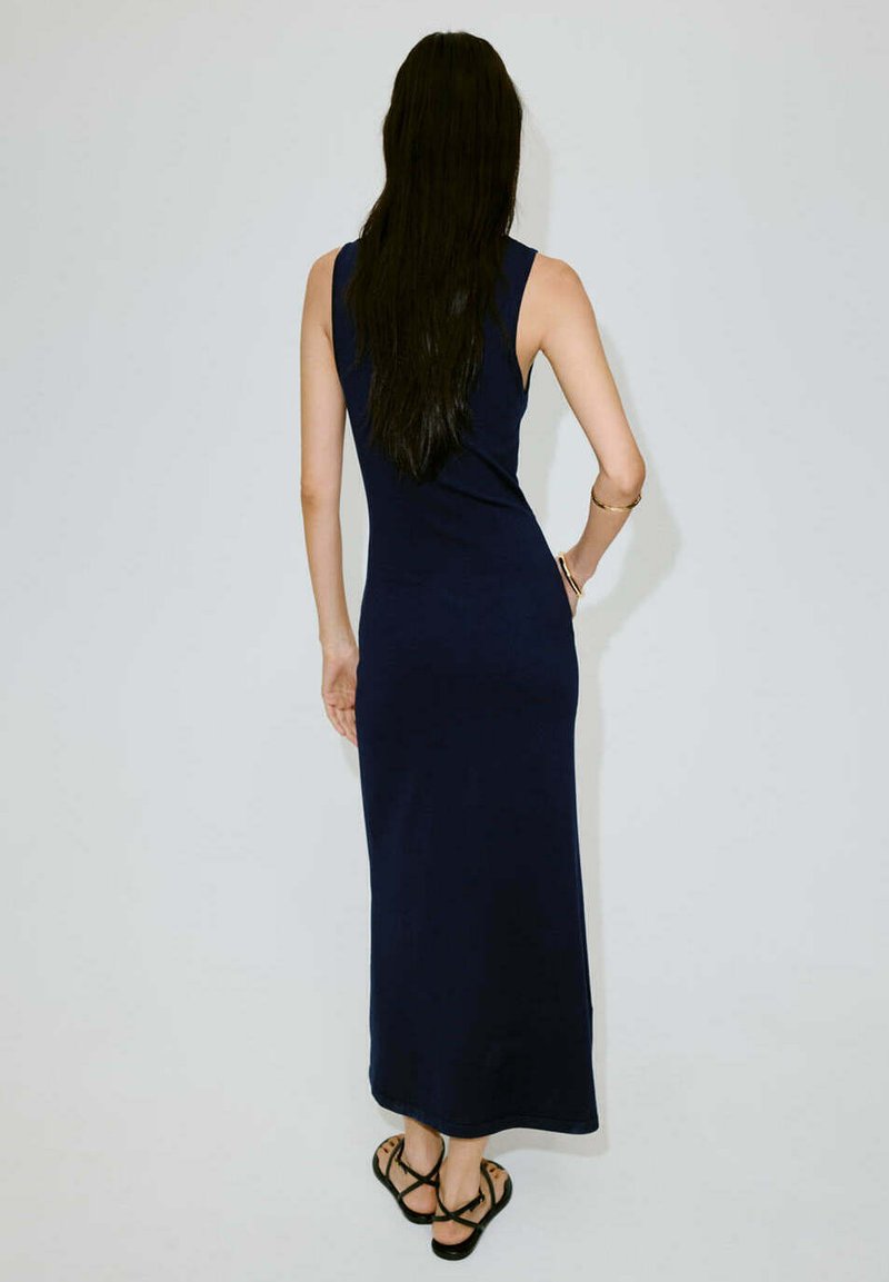 Femme aux longs cheveux foncés portant une robe longue bleu marine sans manches et des sandales noires, debout avec une main sur la hanche, dos tourné vers l'avant.