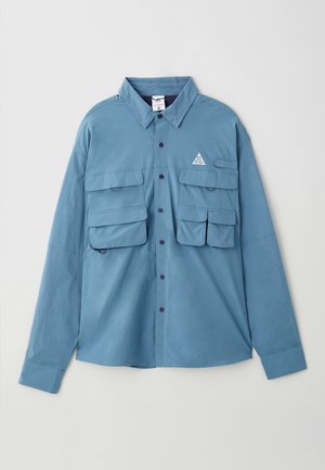 Blå langermet skjorte laget av myk stoff, med to brystlommer, knapplukking, krage og en logo triangulær detalj på brystet.