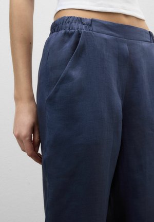 Primo piano di una persona che indossa pantaloni di lino blu scuro con cintura elastica e tasche laterali, abbinati a un top bianco corto.