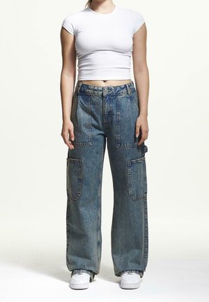 Kobieta w białym obciętym t-shircie, luźnych niebieskich dżinsowych spodniach cargo z wysokim stanem i dużymi kieszeniami oraz białych trampkach, stoi prosto.