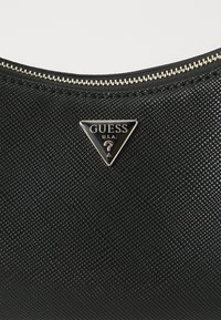 Black textured handbag featuring a triangular metal logo plate embossed with "GUESS U.S.A." and a gold-tone zipper along the top edge.