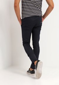 Mężczyzna z dłońmi w kieszeniach, ubrany w czarne dopasowane spodnie, czarne trampki i czarno-białą pasek koszulkę, odchodzi.