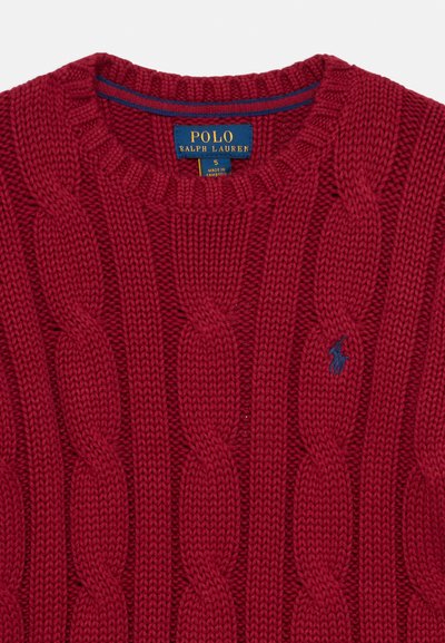 Raudonas megztinis su kabeliniu raštu, turintis apvalią iškirptę, išlankstytomis rankogaliais ir mažą mėlyną logotipą. Pagamintas iš tekstūruoto megztinio audinio su storu raštu.