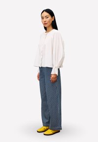 Weißes, langärmeliges Hemd mit gerafftem Vorderteil, kombiniert mit blau-weißen gestreiften weit geschnittenen Hosen und gelben Turnschuhen. Glattes, lässiges Design.