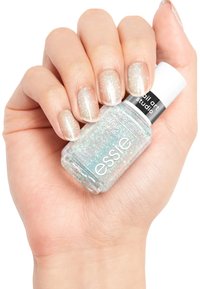 Bouteille de vernis à ongles Essie tenue dans une main ; la bouteille est claire avec un capuchon métallique, contenant un vernis à paillettes scintillantes et iridescentes. Les ongles sont clairs et brillants.