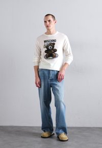 Weißer Pullover mit einem grafischen Motiv eines braunen Teddybären, kombiniert mit hellblauen Jeans und beigen Clogs, vor einem neutralen Hintergrund.