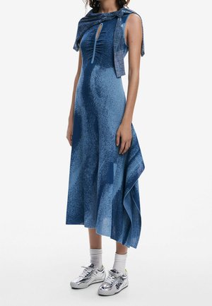 Femme portant une robe midi bleue sans manches avec un ourlet asymétrique, un pull noué sur les épaules, des chaussettes blanches et des baskets argentées.