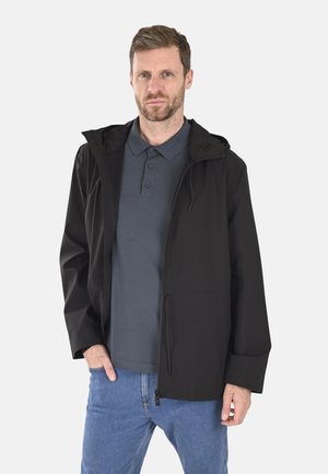 Veste noire légère avec capuche, dotée d'une fermeture éclair à l'avant, de poches latérales et d'une finition lisse, portée par-dessus un polo gris et un jean bleu.