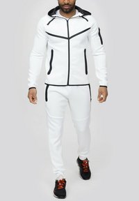 Ensemble de sport blanc avec des accents noirs, comprenant une veste zippée avec capuche et poches zippées, associée à un pantalon fuselé.