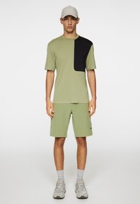 Vihreä urheilupaita, jossa on musta paneeli, yhdistettynä samanvärisiin shortseihin. Päällä harmaat lenkkarit ja vaaleanharmaa lippis.