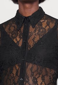 Chemisier en dentelle noire avec un col, présentant des motifs floraux et des sections transparentes, fermé par de petits boutons noirs.