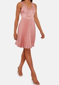 Rosa plisserad klänning med V-ringning, genomskinliga detaljer och blomstermönster på bysten. Bärs med klara ankelsandaler.