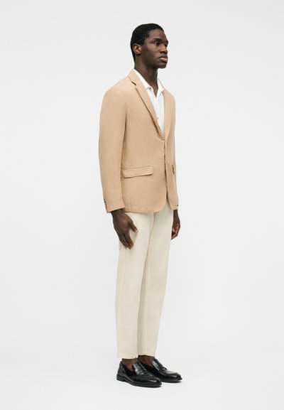 Άνδρας όρθιος, φοράει μπεζ σακάκι, άσπρο πουκάμισο, κρεμ παντελόνι και μαύρα μοκασίνια, σε απλό λευκό φόντο.