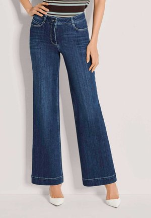 Frau trägt hoch taillierte dunkelblaue weit geschnittene Jeans mit weißen, spitzen Absätzen und einem gestreiften Oberteil, steht vor einer einfarbigen Wand.