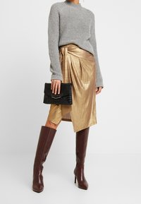 Grå stickad tröja, gyllene metallisk kjol med veck, svart texturerad clutch och bruna knähöga klackstövlar. Släta material och lagerdesign.