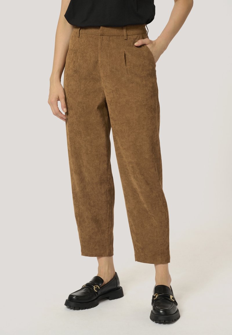 Pantalon en velours côtelé marron taille haute, fuselé, porté avec des mocassins noirs, montré de la taille aux pieds sur un fond clair.