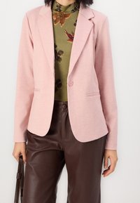 Světle růžový blazer s klopovými límci, jedním knoflíkem a dvěma bočními kapsami, kombinovaný se zeleným květinovým topem a hnědými kalhotami z umělé kůže.
