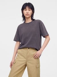 Personne aux cheveux noirs mi-longs porte un t-shirt ample gris foncé et un pantalon cargo beige, debout avec une main sur la hanche.