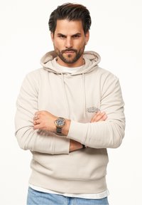 Beige hoodie with a drawstring hood, ribbed cuffs, and a logo on the chest. Worn over a white shirt with light blue jeans and a black watch.