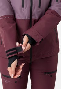 Giacca da sci bordeaux con sezione superiore viola, dotata di tasche con zip, polsini regolabili e dettagli con logo, realizzata in materiale resistente all'acqua.