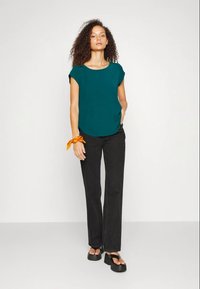 Top teal a maniche corte con scollatura rotonda, abbinato a pantaloni neri a gamba dritta e sandali neri chunky. Accessorio arancione al polso.
