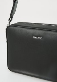 Black rectangular crossbody bag made of textured synthetic material, featuring a silver zipper and a prominent Calvin Klein logo.