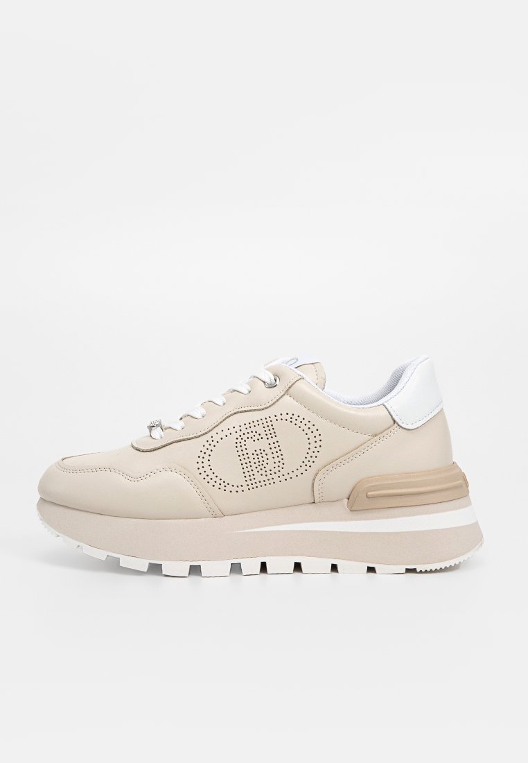 Zapatilla beige con diseño perforado, acentos blancos, suela texturizada y detalle de logo. Presenta una entresuela acolchada y cierre de cordones.