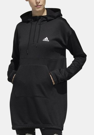 Donna che indossa un vestito con cappuccio nero Adidas con tasca frontale e cappuccio, mani nelle tasche, mostra il logo Adidas bianco sul petto.