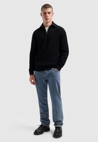 Jongeman in een zwart half-zip trui, lichtblauwe jeans en zwarte leren schoenen, staand met één hand in zijn zak op een grijze achtergrond.