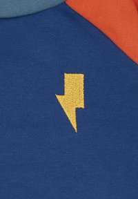 Jersey di cotone blu con ricamo di un fulmine giallo. Include maniche raglan turchesi e arancioni, texture liscia e colori vivaci.