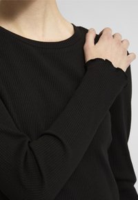 Top negro de manga larga con textura acanalada, con un cuello redondo y puños suavemente festoneados, destacando su superficie texturizada contra un fondo liso.