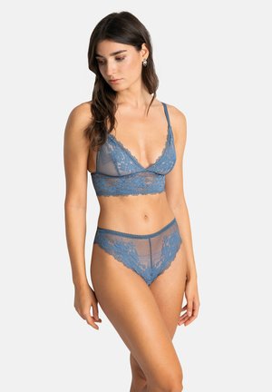 Femme aux longs cheveux bruns portant un bralette en dentelle bleue et un slip assorti, posant devant un fond clair uni.
