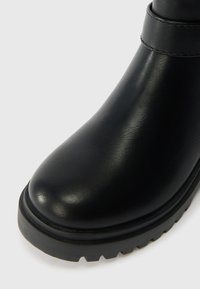 Μαύρη δερμάτινη μποτάκι αστραγάλου με στρογγυλή μύτη, καουτσούκ σόλα και ραφές σε λείο επιφάνεια. Ο σχεδιασμός είναι μινιμαλιστικός και ανθεκτικός.